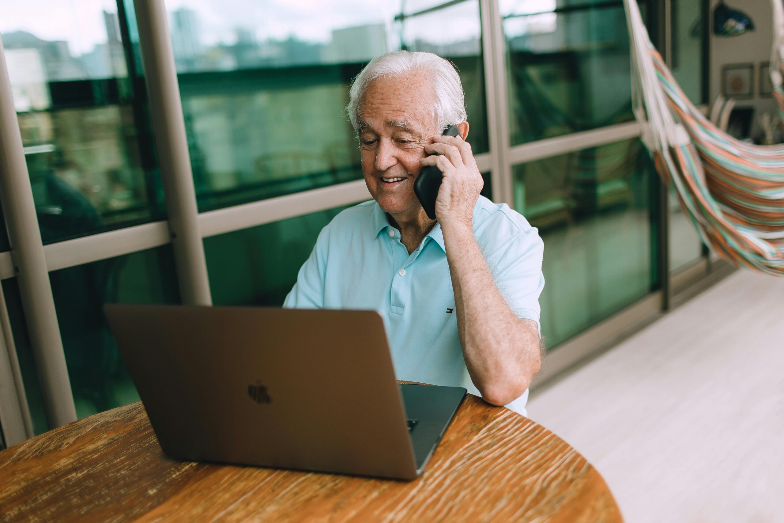 A senior man talking on phone while using a laptop at home, wearing a casual blue shirt.